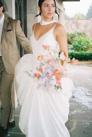 A romantic wedding scene showing details of a flowing white dress and pastel pink and purple floral bouquet.