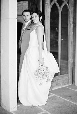Black and white wedding photo showing bride in elegant white dress holding bouquet and groom in suit standing by arched doorway.