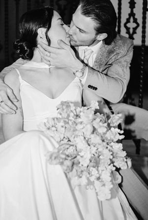 Black and white wedding photo shows couple sharing a romantic kiss while holding an elegant white floral bouquet.