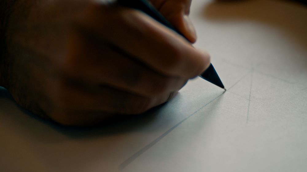 A close-up view shows someone's hand writing with a pen on white paper.