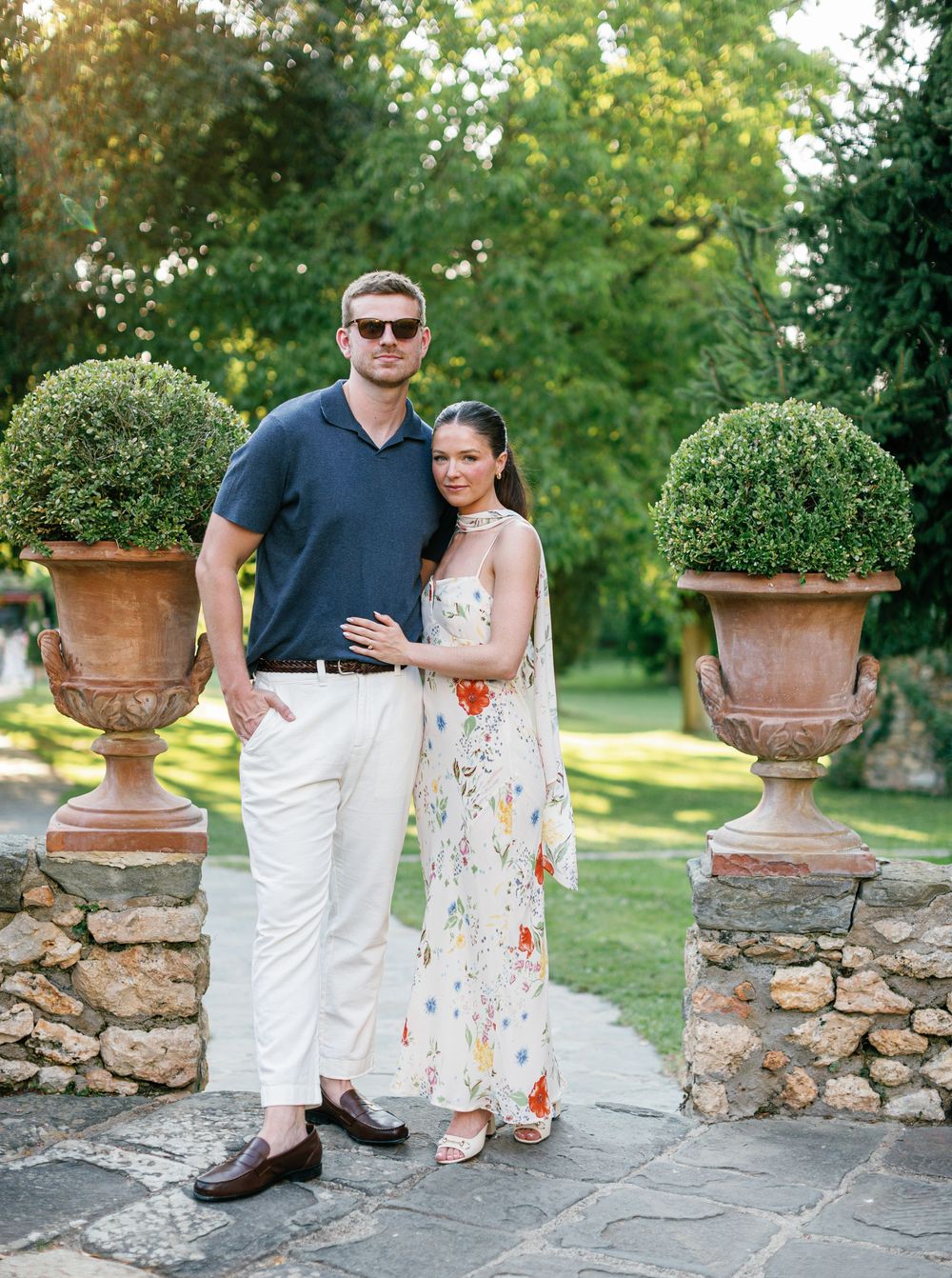 A couple stands together at a stone wall with large terracotta urns and lush greenery in the background at Borgo San Luigi.