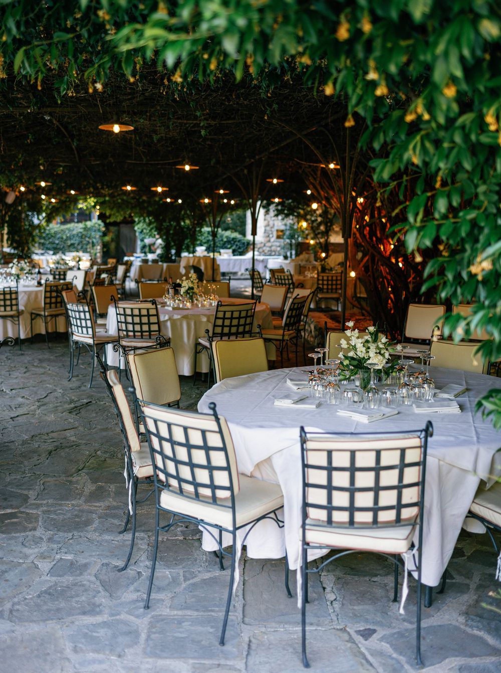 Elegant outdoor wedding reception setup with white tablecloths, grid-backed chairs, and lush greenery overhead at Borgo San Luigi.