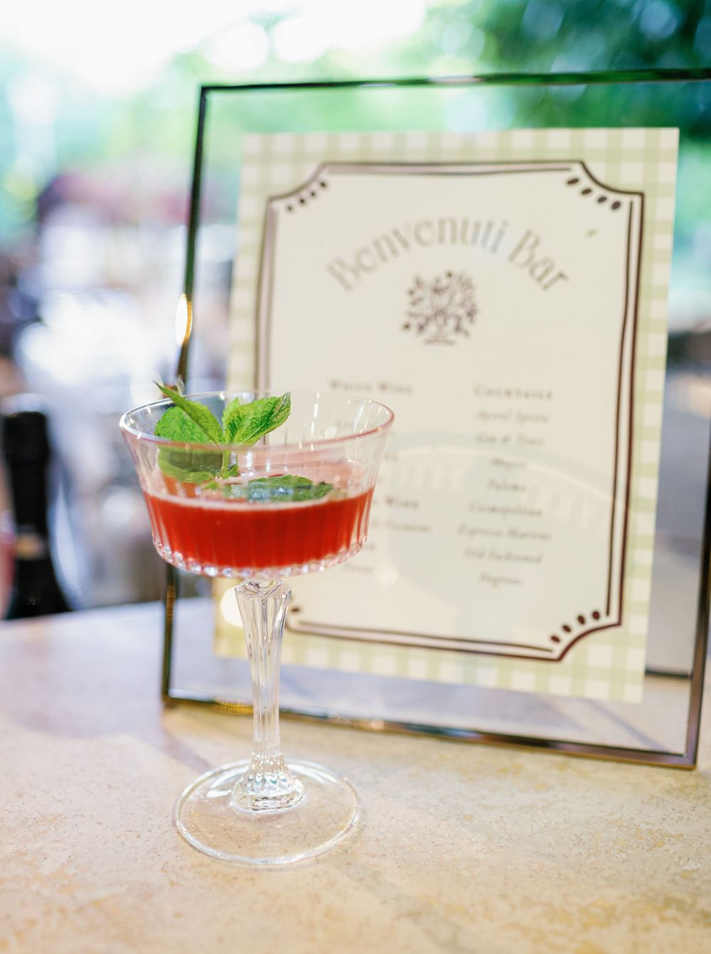 A row of elegant cocktail glasses with red drinks and fresh mint garnishes sit beside vintage-style menu cards at a bar.