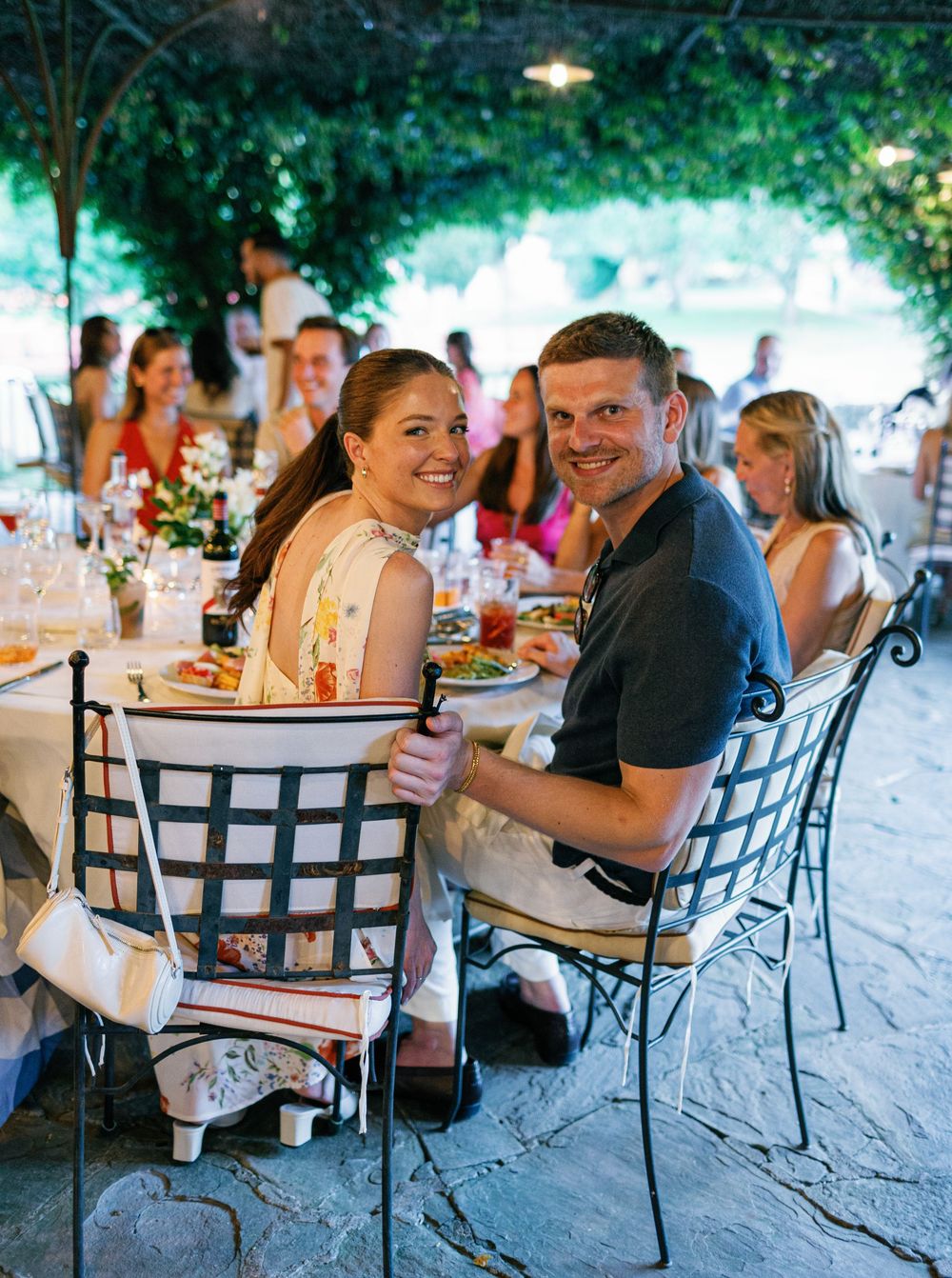 Wedding guests enjoy an outdoor dining reception under a canopy of greenery at elegant tables and chairs.