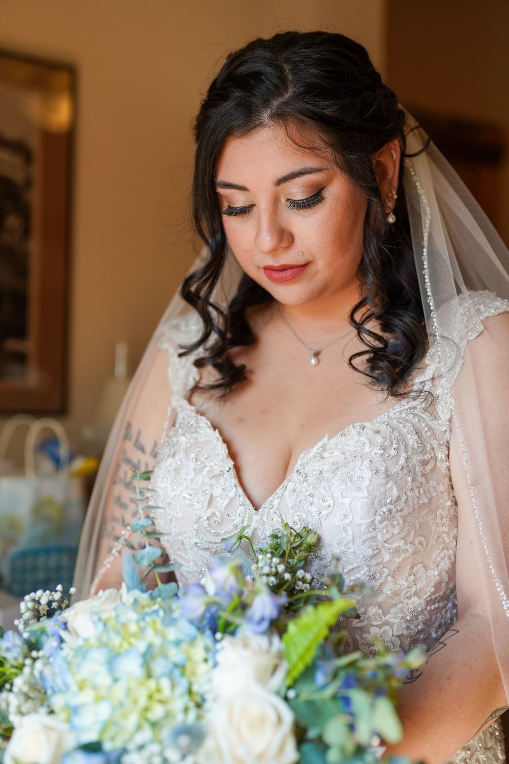 Bride in lace wedding dress holds a blue and white bouquet while looking down in a warm-lit room.