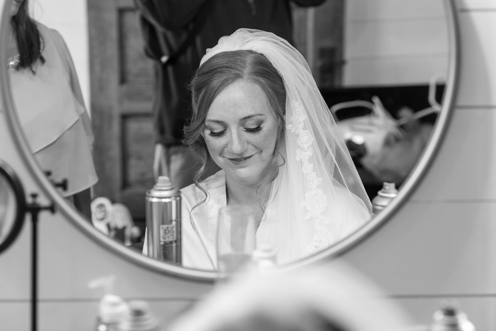 A bride in a white wedding gown and veil smiles while getting ready in front of round mirrors before the ceremony.