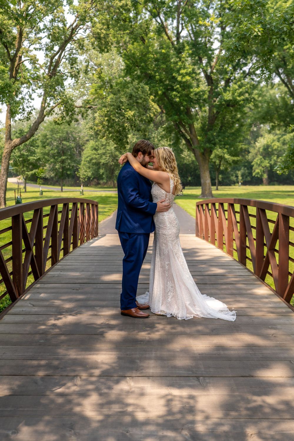 A romantic moment captured on a wooden bridge surrounded by lush green trees in a park setting.