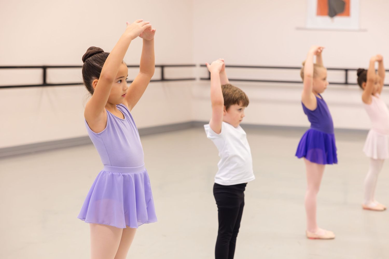 Young students practice ballet positions with raised arms in a dance studio with a barre and mirrors.