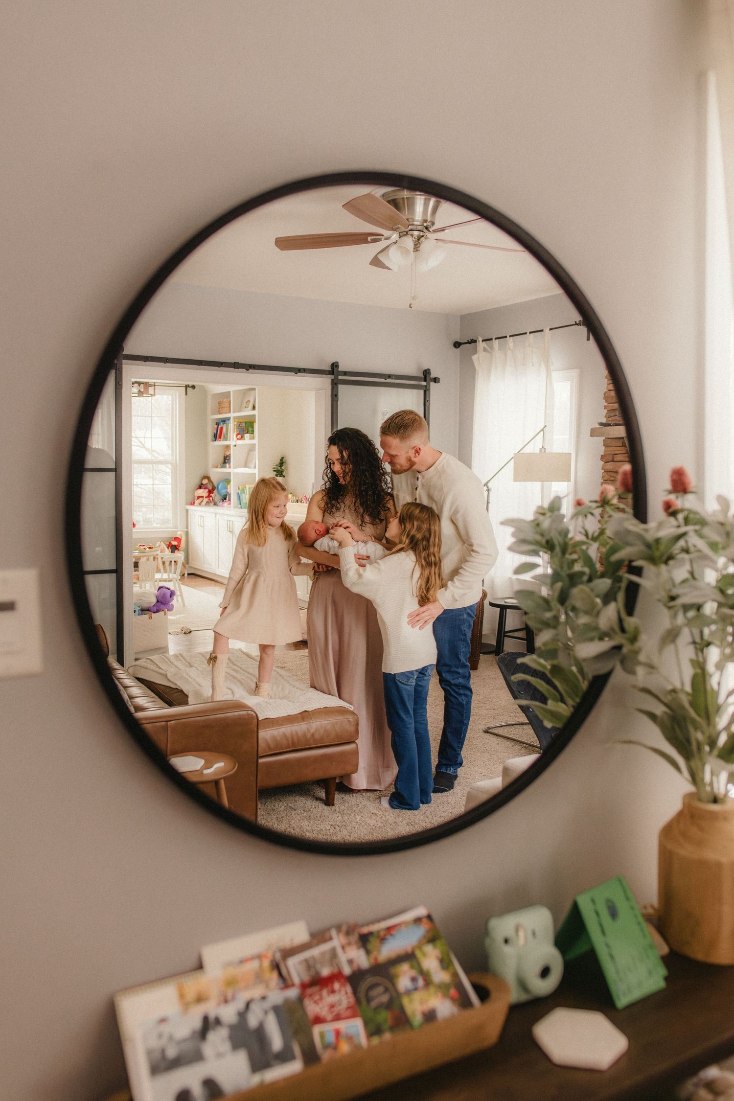 A round mirror reflects a family gathered in a cozy bedroom with neutral decor and natural light streaming through windows.