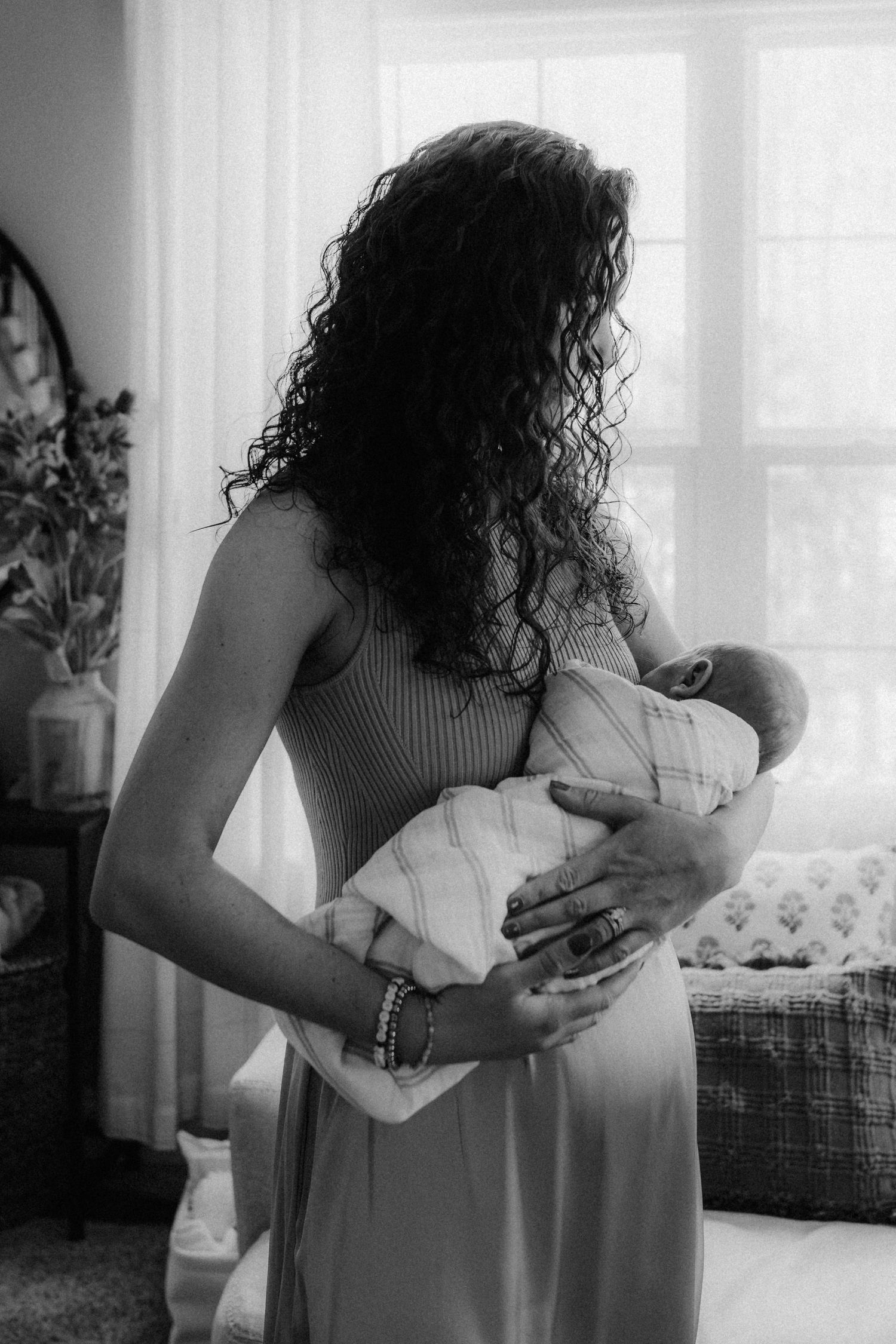 A black and white photograph shows a tender moment between a parent cradling a swaddled newborn baby by a window.