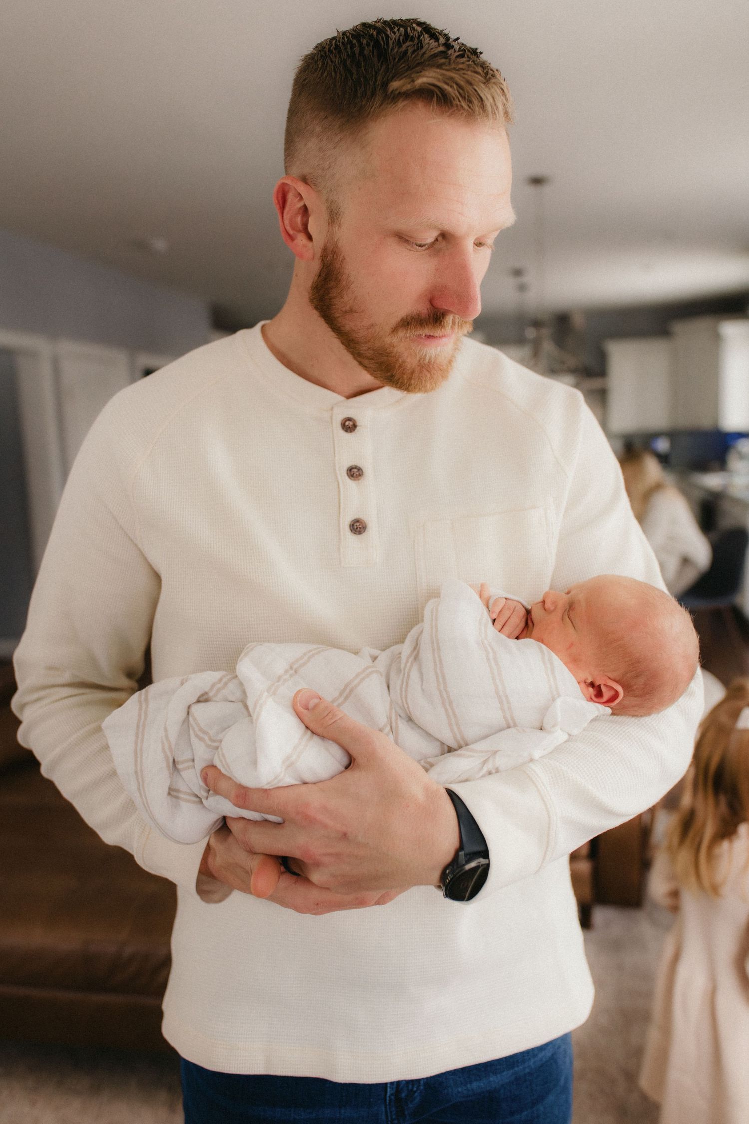 Person in white henley sweater tenderly holding a swaddled newborn baby indoors.