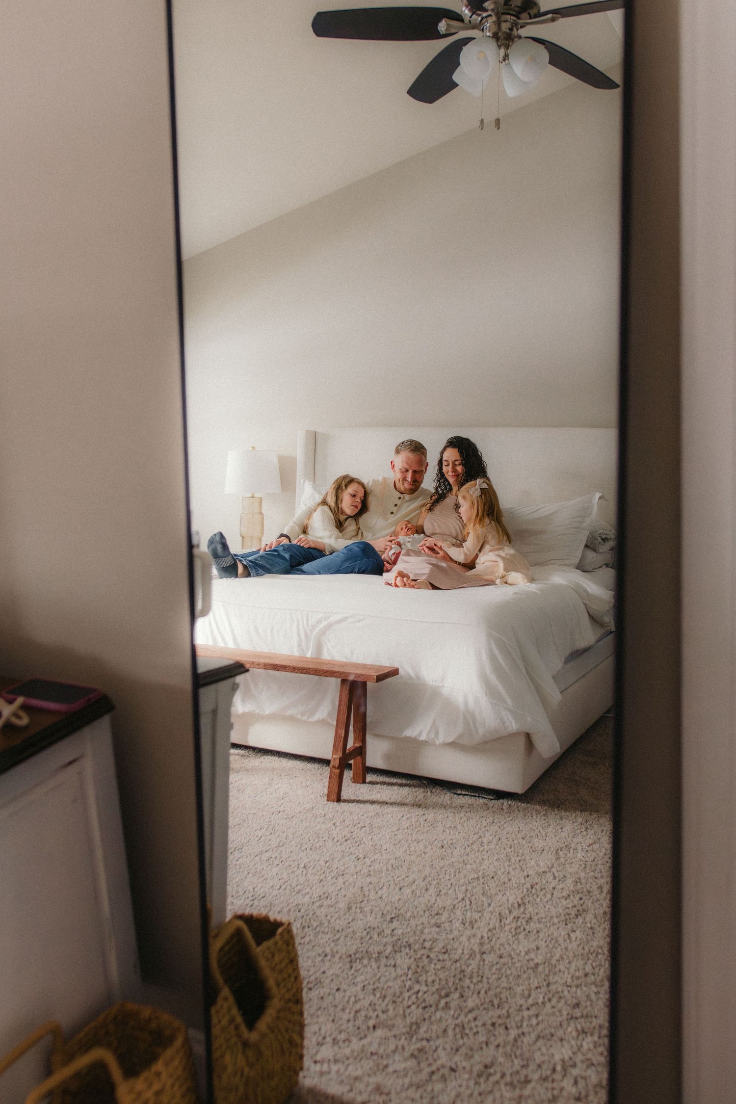 A mirror reflection shows a family relaxing together on a white bed in a bright, minimalist bedroom.