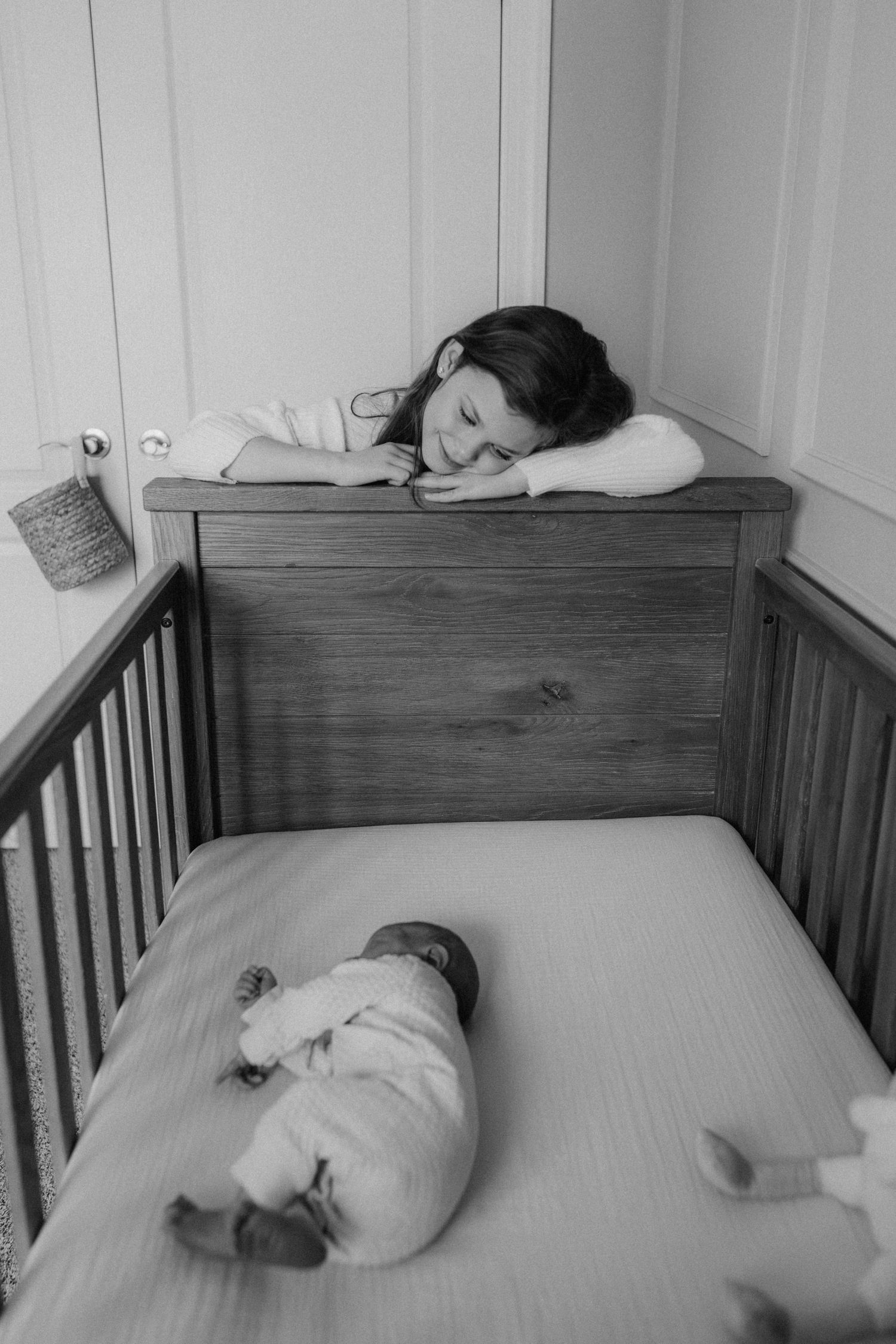 A black and white photo showing someone resting on a wooden crib while a baby sleeps inside.