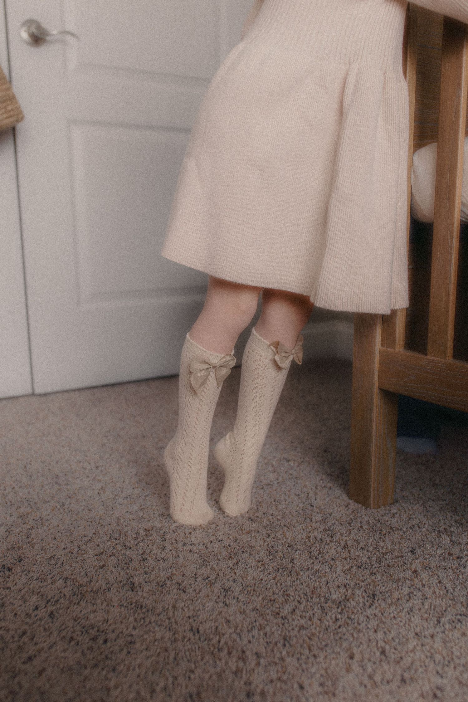 A person wearing a beige skirt and cream-colored knit socks with bows stands on gray carpet near a wooden chair leg.