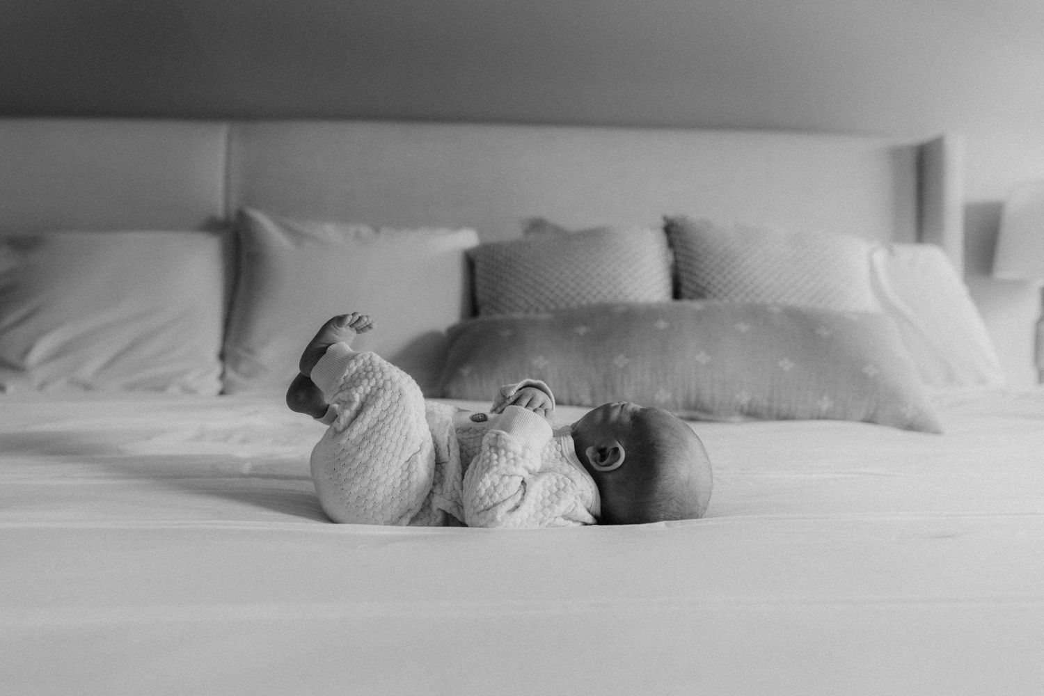 A baby lies on a white bed wearing light-colored clothing in a peaceful black and white photograph.