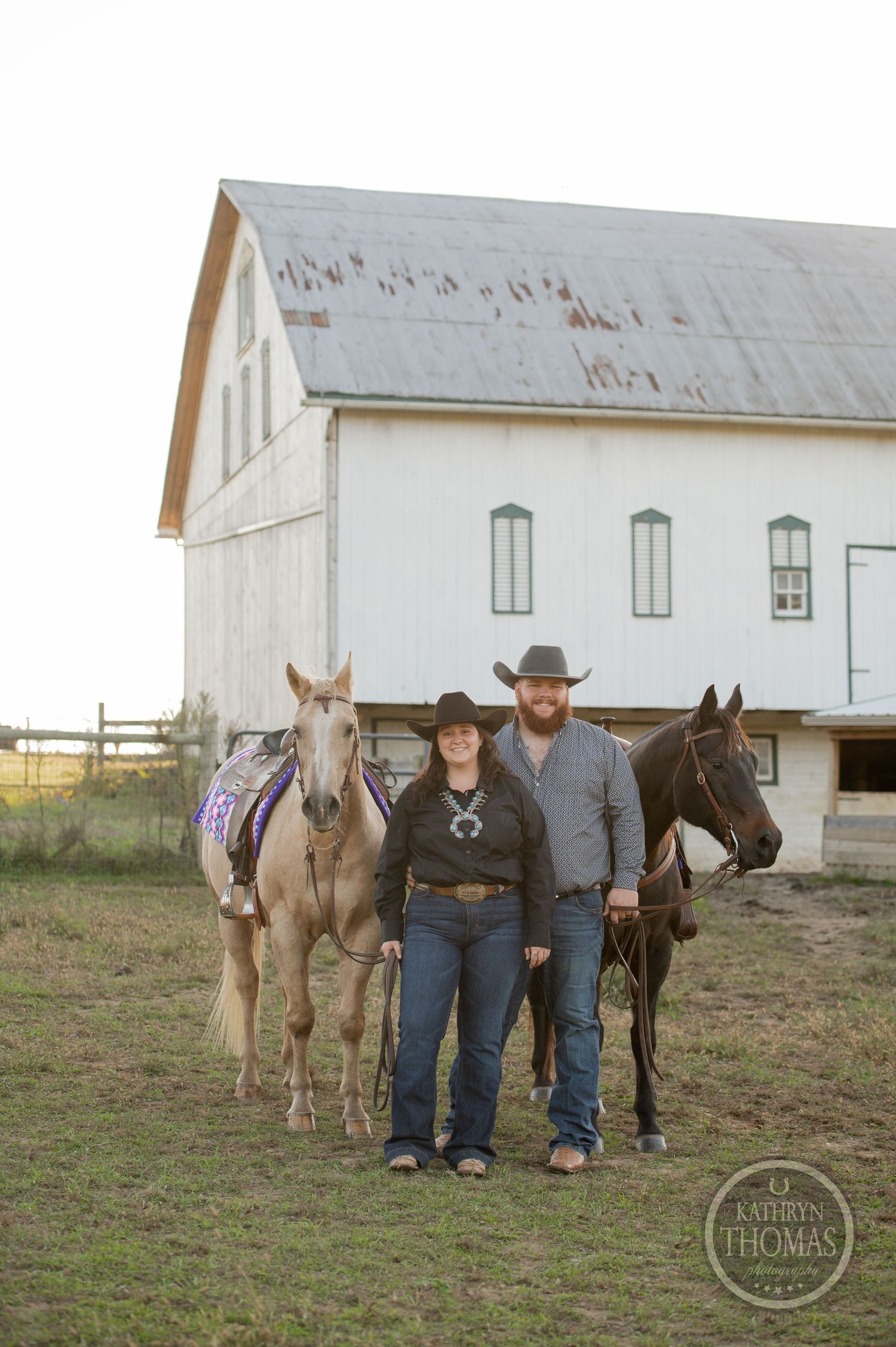 Two people stand between palomino and dark horses in front of a classic white barn.