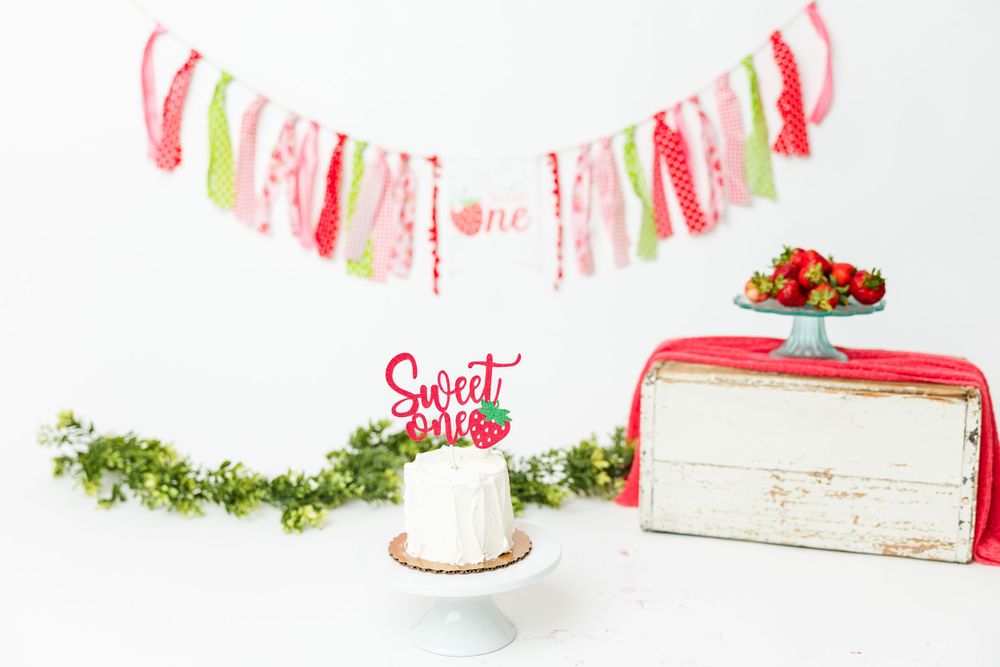 Small white birthday cake with pink topper displayed next to vintage red bread box under festive pink and green banner.