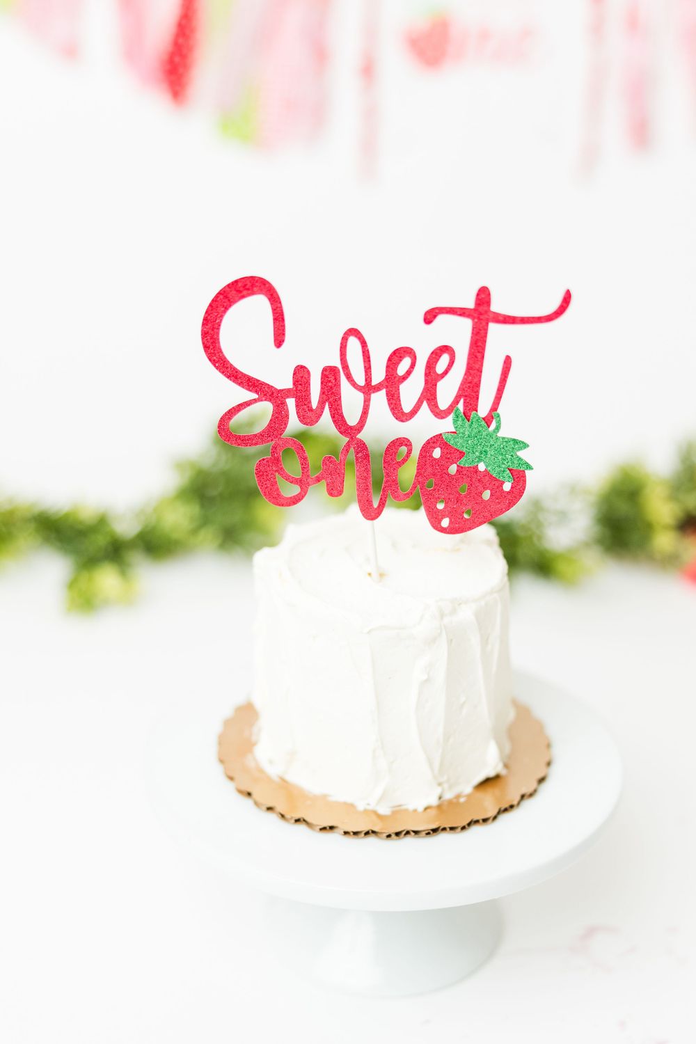 A white frosted mini cake with a red glittery 'Sweet' cake topper and strawberry design on a small cake stand.