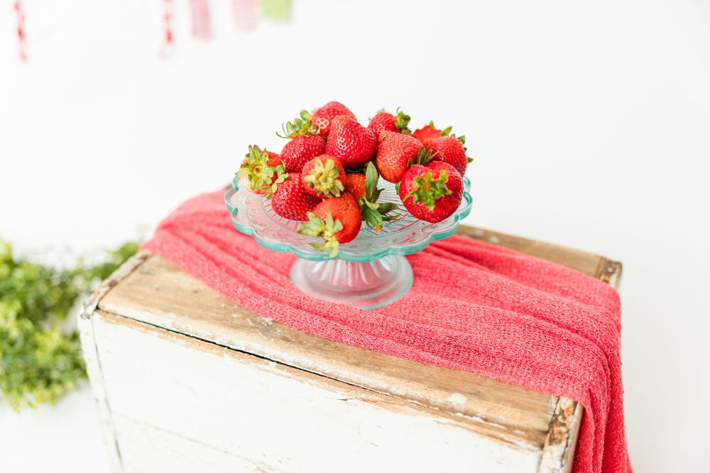 Fresh red strawberries arranged in a glass pedestal bowl on pink towel against white background.