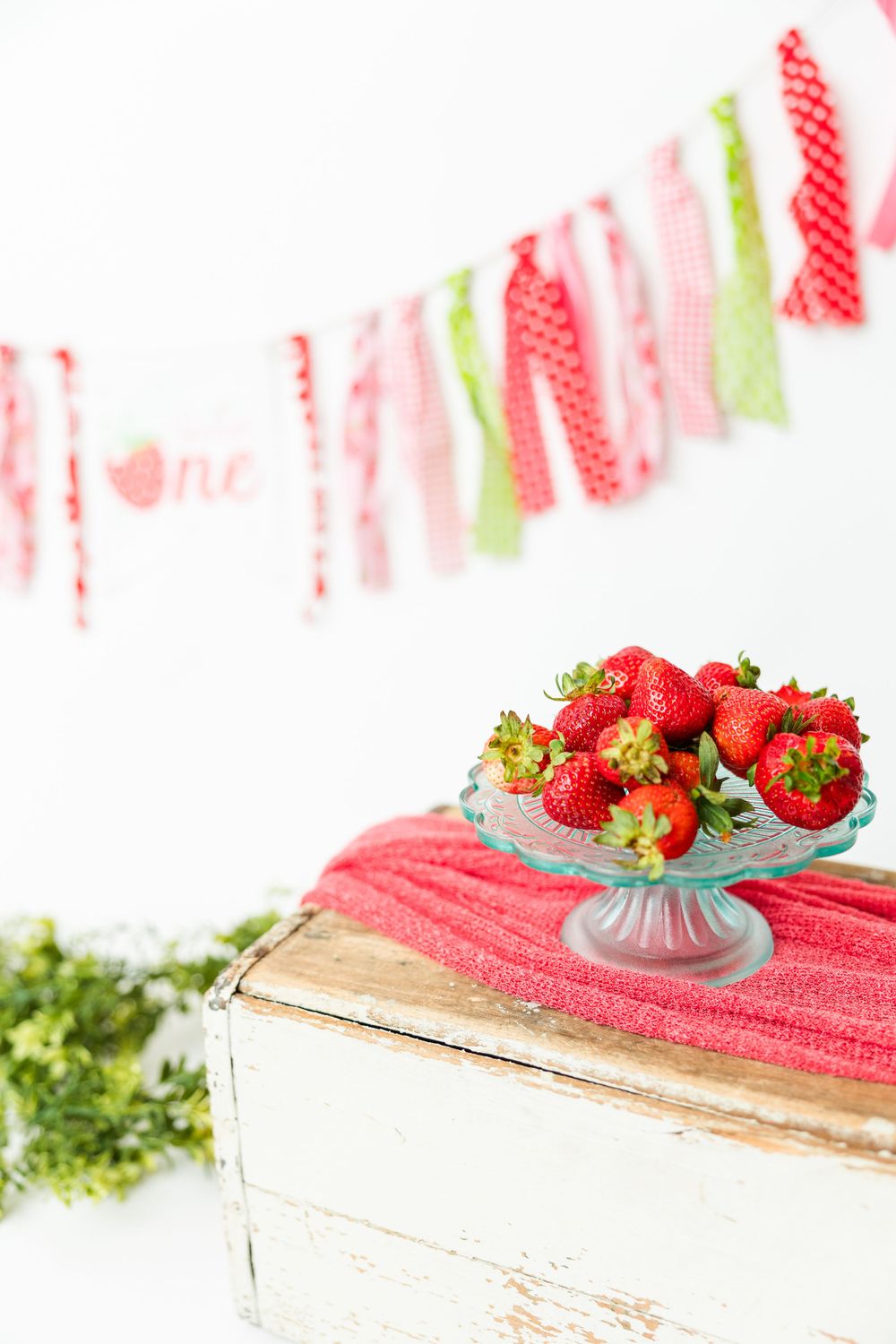 Fresh strawberries displayed in a vintage glass compote on top of a white rustic wooden box with decorative ribbon banner.