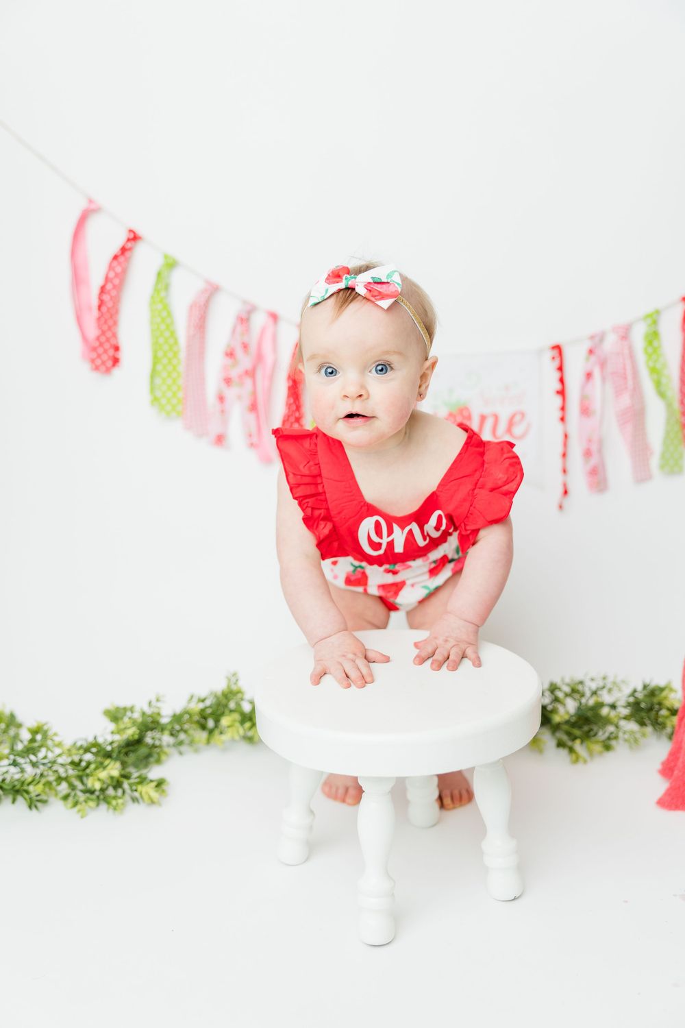 Baby wearing red 'One' outfit sits on white stool with pink and green banner decorations for first birthday photoshoot.