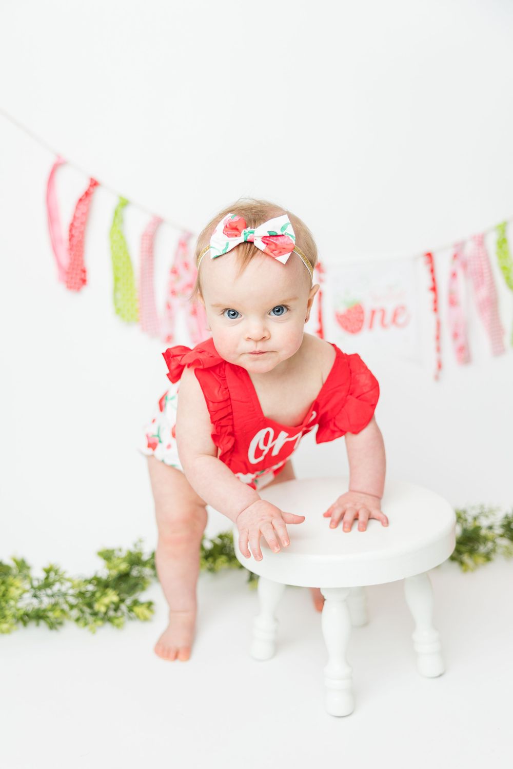 Young child in red ruffled outfit stands on white stool with pink and green banner decorations in background.