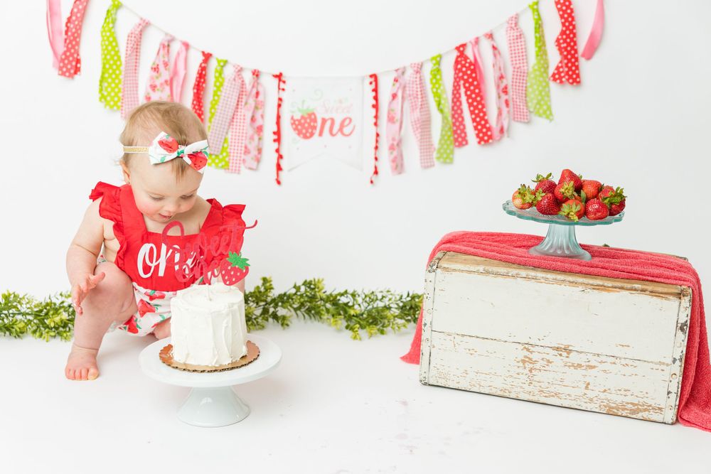 A festive cake smash setup with a white cake, colorful tassel banner, and rustic wooden box decorated with strawberries.