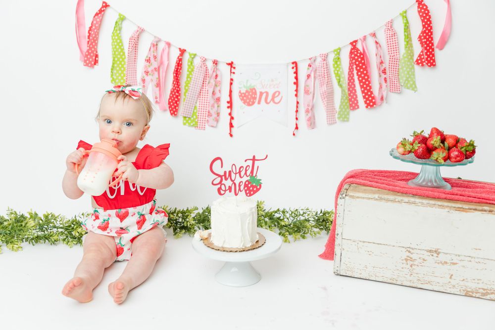 Sweet One birthday setup with pink and mint decorative ribbon banner, cake, and fresh strawberries on white background.