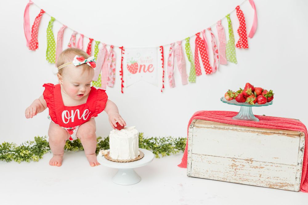 First birthday celebration setup with vintage pink bread box, cake smash decorations, and strawberries on white backdrop.