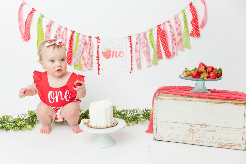 A pink and gold first birthday setup with a smash cake, decorative banner, and vintage white bread box.