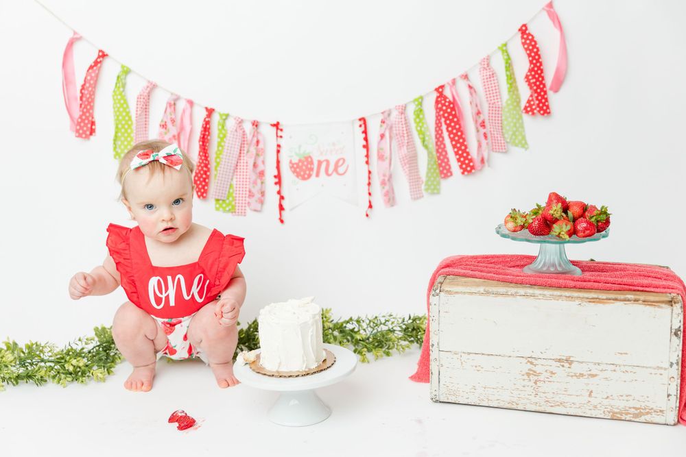 A cake smash photo setup with red and pink decor, a vintage bread box, and greenery garland against white backdrop.