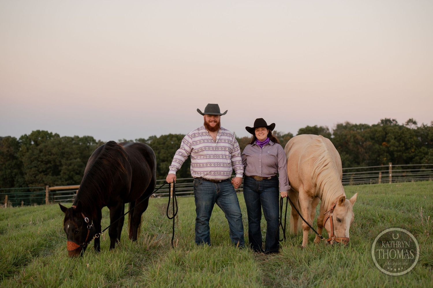 Two people in cowboy attire stand between black and cream-colored horses in a fenced field at dusk.