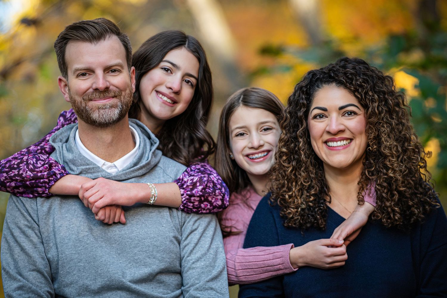 A family of three sits together on a front porch step, dressed in casual fall attire with a decorative wreath on the door.