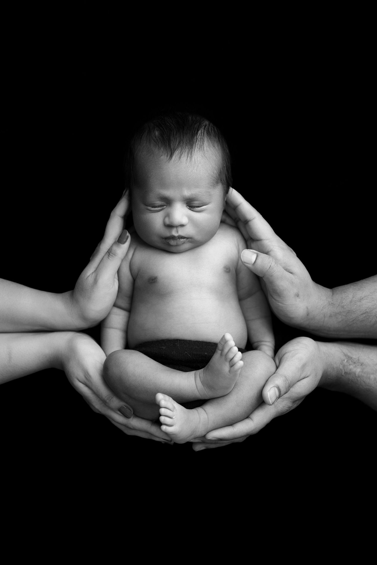 A newborn baby lies peacefully in supporting hands against a black background in a dramatic black and white portrait.