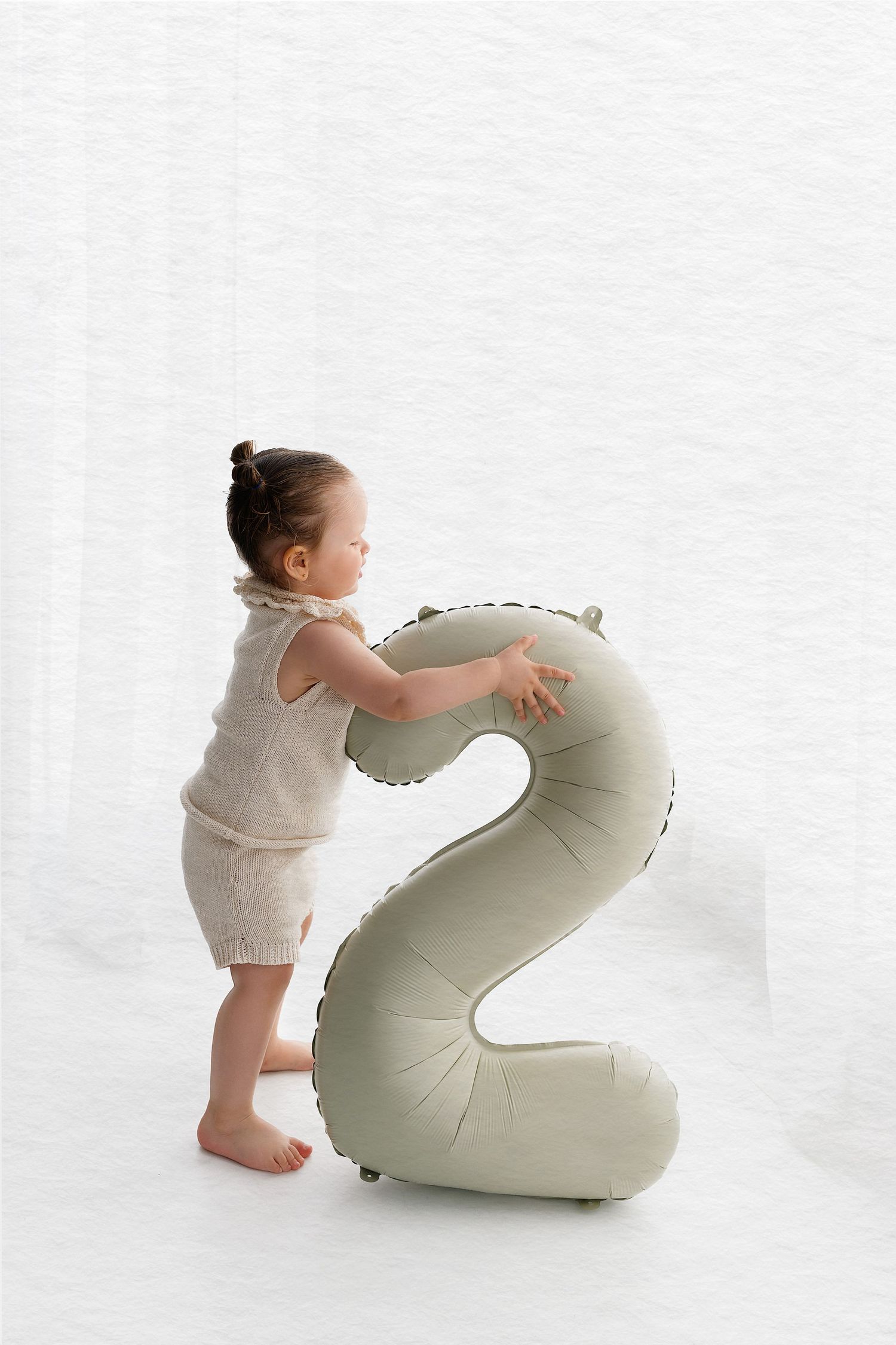 A small child in beige clothing stands next to a large decorative number 2 pillow on a white background.