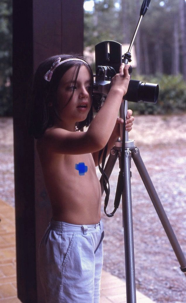Une Fille avec un serre tête rose regarde dans le viseur d'un ancien appareil photo argentique et ajuste la mise au point avec sa main