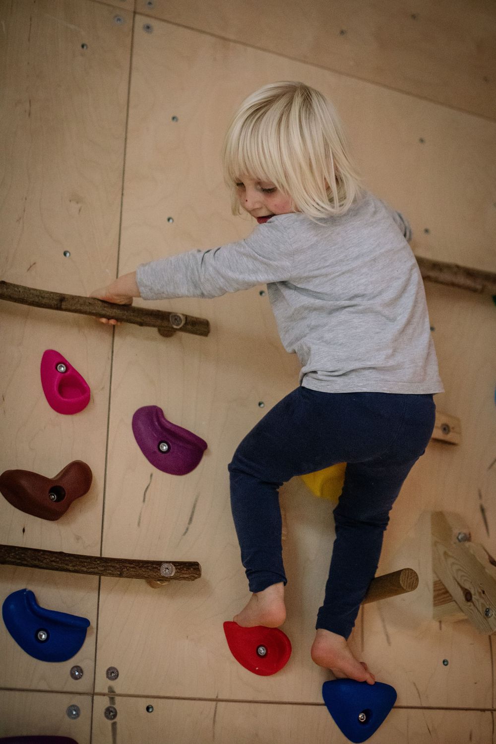 A child climbing on an indoor rock climbing wall with colorful holds wearing gray and blue clothing.