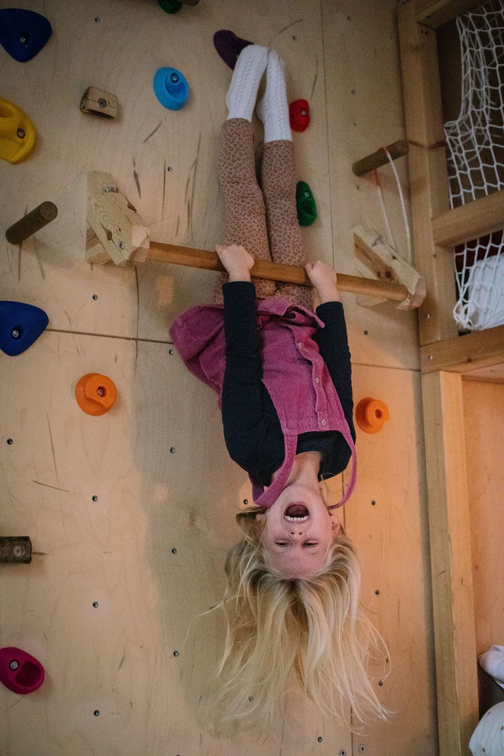 Someone climbs upside down on an indoor rock climbing wall with colored climbing holds and hangs playfully with hair down.