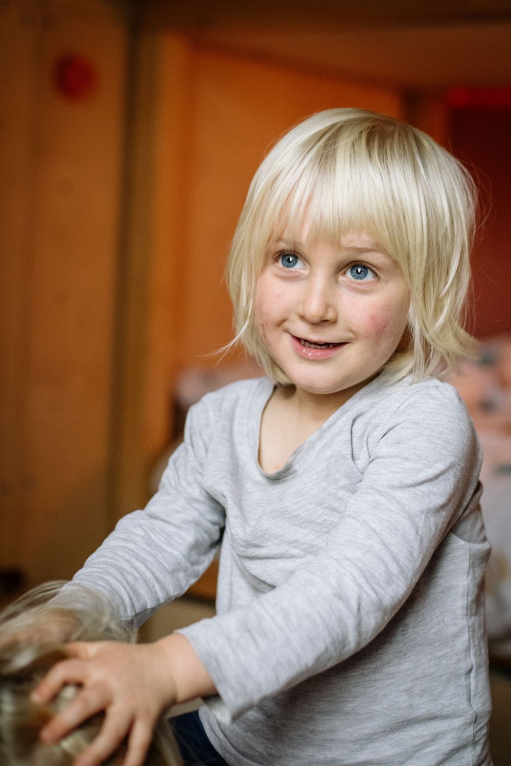 Young child with light blonde hair and bright blue eyes wearing a gray long-sleeve shirt smiles warmly at the camera.