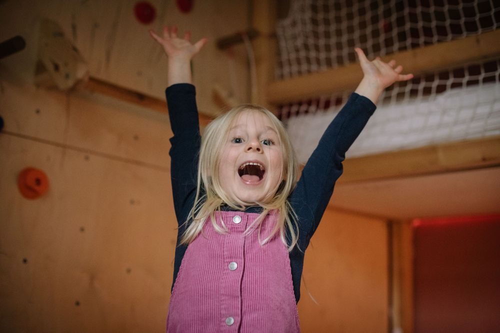 A young child in a pink dress raises their arms with pure joy and excitement against an indoor backdrop.