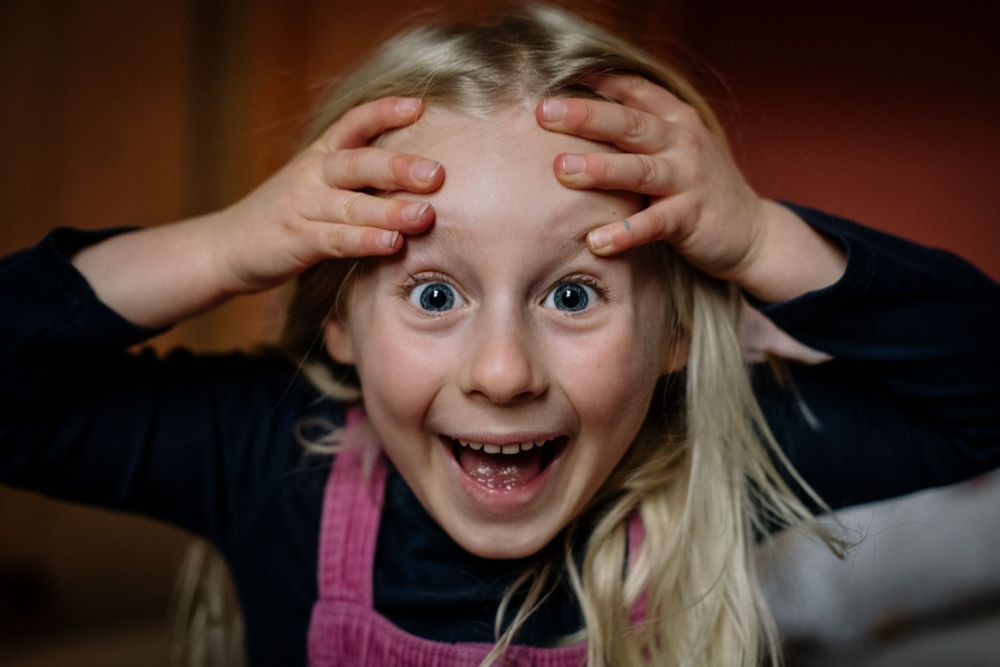 A blonde child with wide eyes makes an excited expression while pressing hands against their forehead.