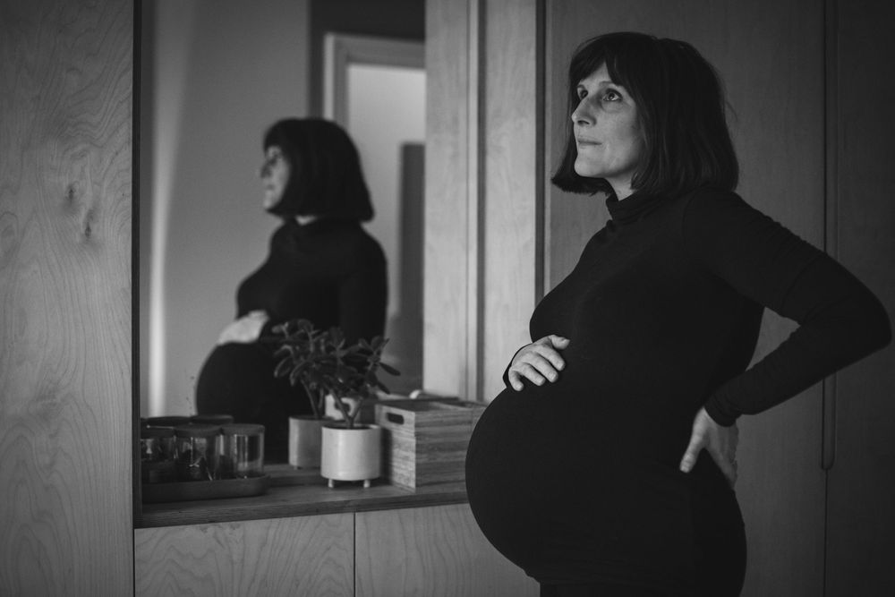 Black and white photo showing reflection in mirror of pregnant person wearing dark clothing with hands on hips.