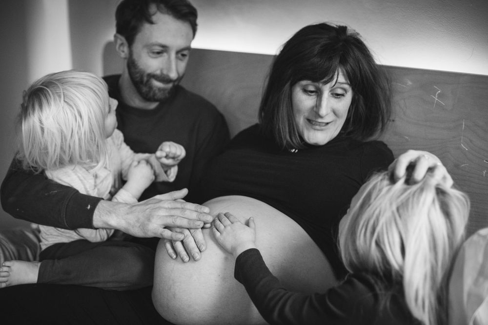 Black and white photograph showing a pregnant woman's belly being touched by two people in an intimate family moment.