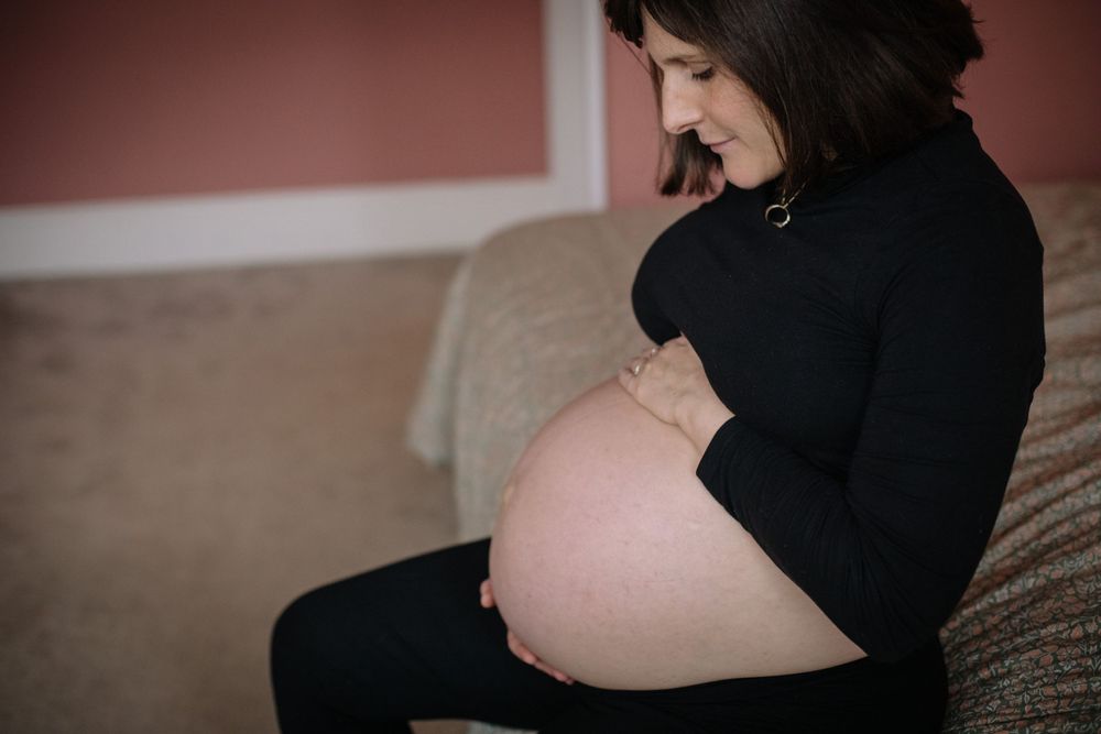 Side view of an expectant mother in a black sweater sitting on a couch and lovingly touching her pregnant belly.