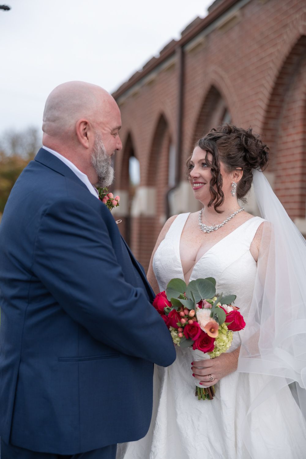 A wedding couple shares a tender moment against a brick building backdrop with the bride holding a red and pink bouquet.