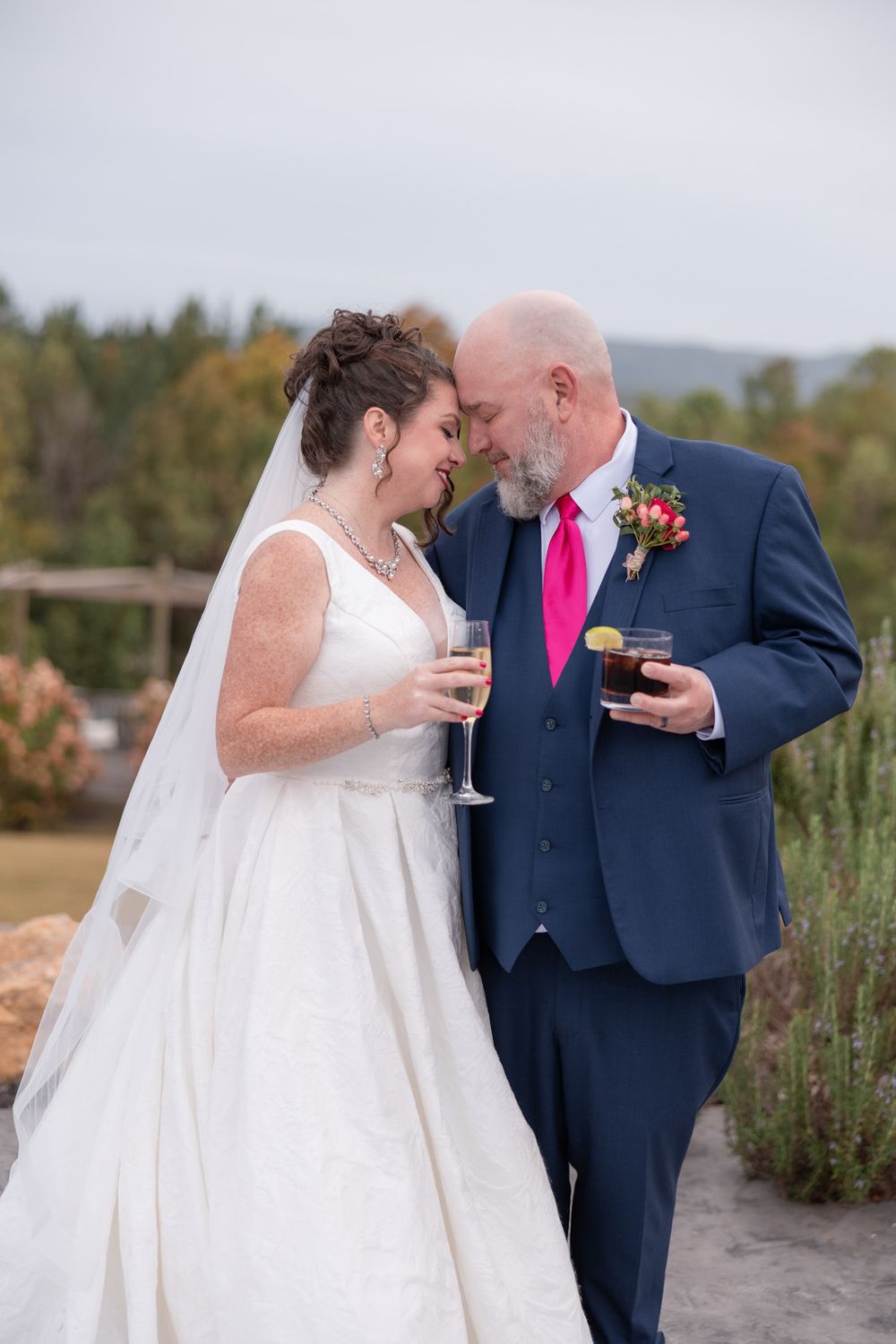 A bride and groom share an intimate moment while holding wine glasses at an outdoor wedding venue with autumn scenery.