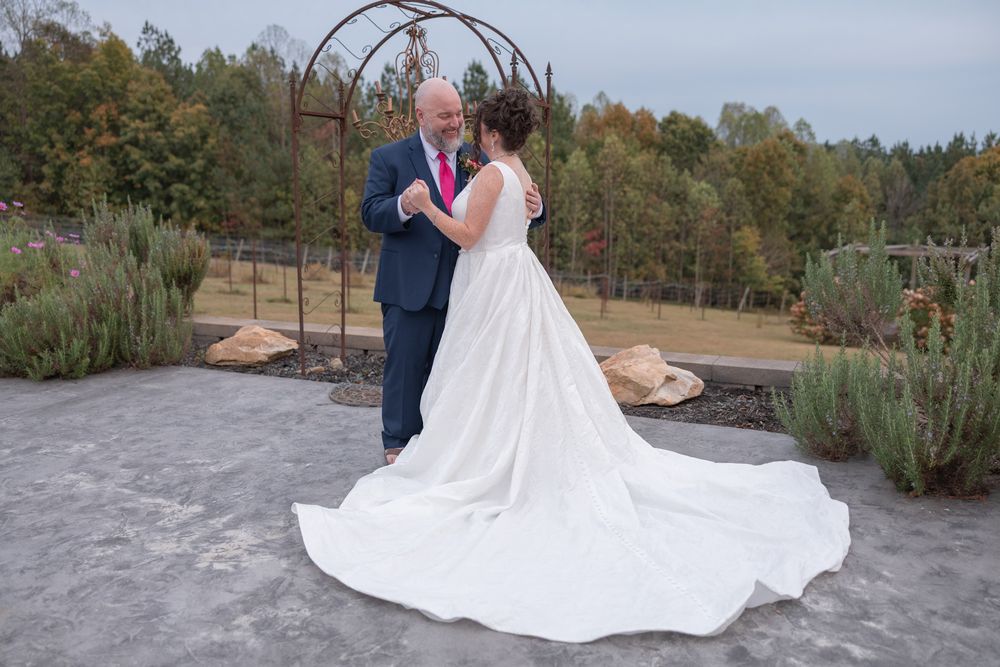 A couple in wedding attire embraces on a sandy beach surrounded by pine trees and coastal vegetation.