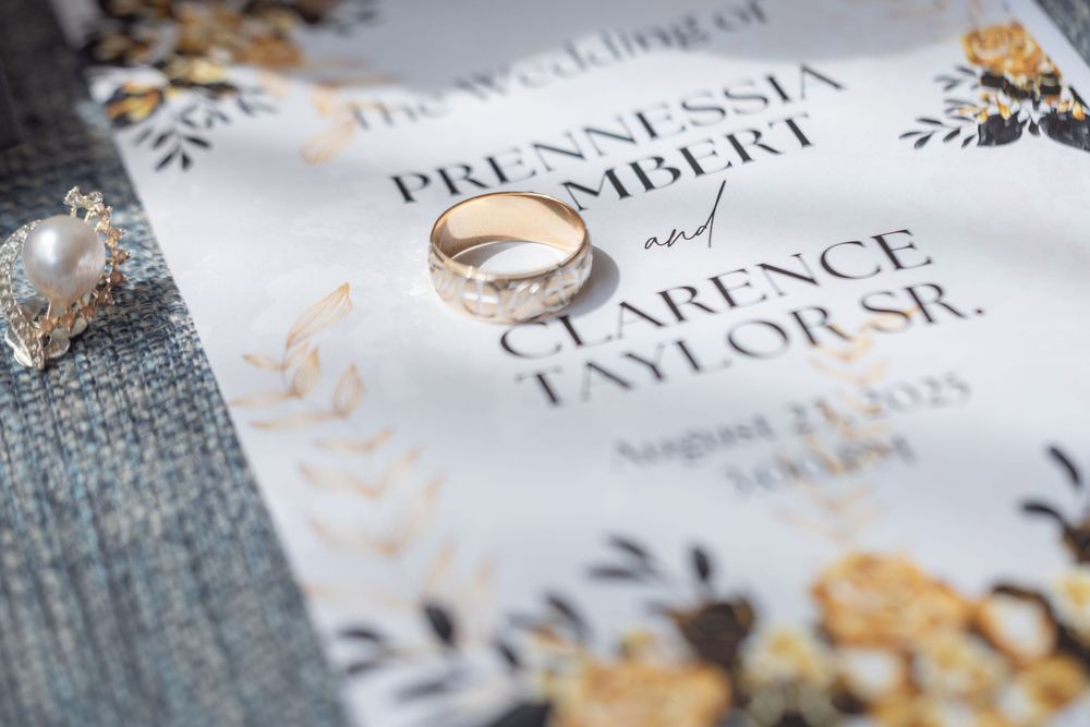 Wedding rings rest on an elegant invitation with floral designs and gold accents against a textured gray background.