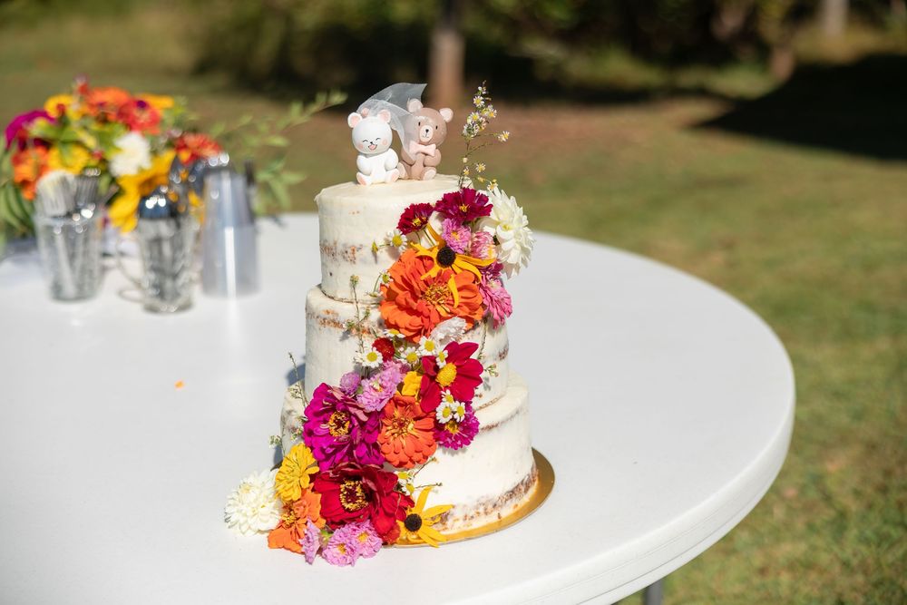 A three-tiered white wedding cake decorated with vibrant pink, orange and yellow flowers sits on an outdoor table.