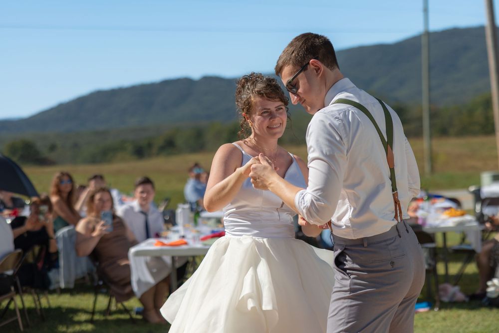 A couple shares their first dance at an outdoor wedding reception with mountains and guests in the background.